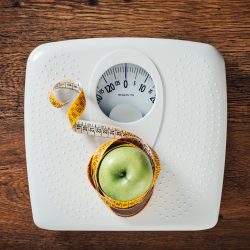 Green apple wrapped in a tape measure on a white scale, wooden surface on background, dieting and weight loss concept