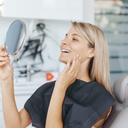 Young woman patient is looking in the mirror and admires her new smile after dental treatment in the dentistry clinic.