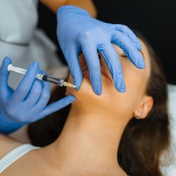 Cosmetician in gloves gives chin botox injection to female patient on treatment table. Rejuvenation procedure in beautician salon. Doctor with syringe and woman, cosmetic surgery against wrinkles